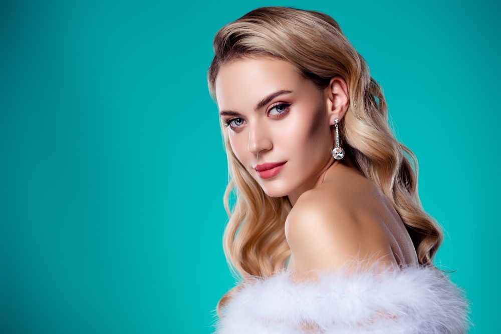 Glamorous woman with wavy blonde hair and crystal drop earring against teal backdrop, wrapped in white faux fur.