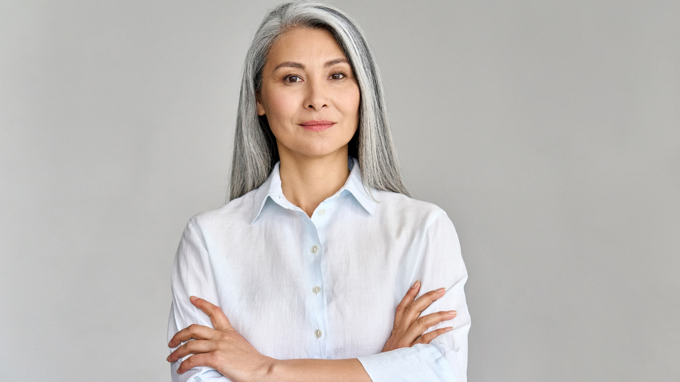 A confident woman with long, beautifully blended silver hair stands against a soft grey background. Her sleek, natural look reflects expert grey blending techniques—showcasing the elegant, seamless results available at Honeys Hair & Beauty in Taunton.