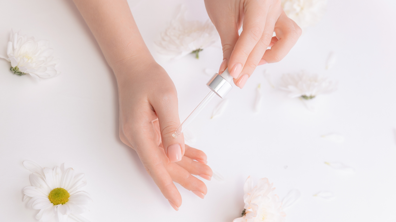 A close-up of a hand applying nourishing cuticle oil with a dropper onto freshly manicured nails, surrounded by soft white daisies. The image reflects gentle at-home nail care, perfect for maintaining nail infills between professional salon visits at Honeys Hair & Beauty in Taunton.