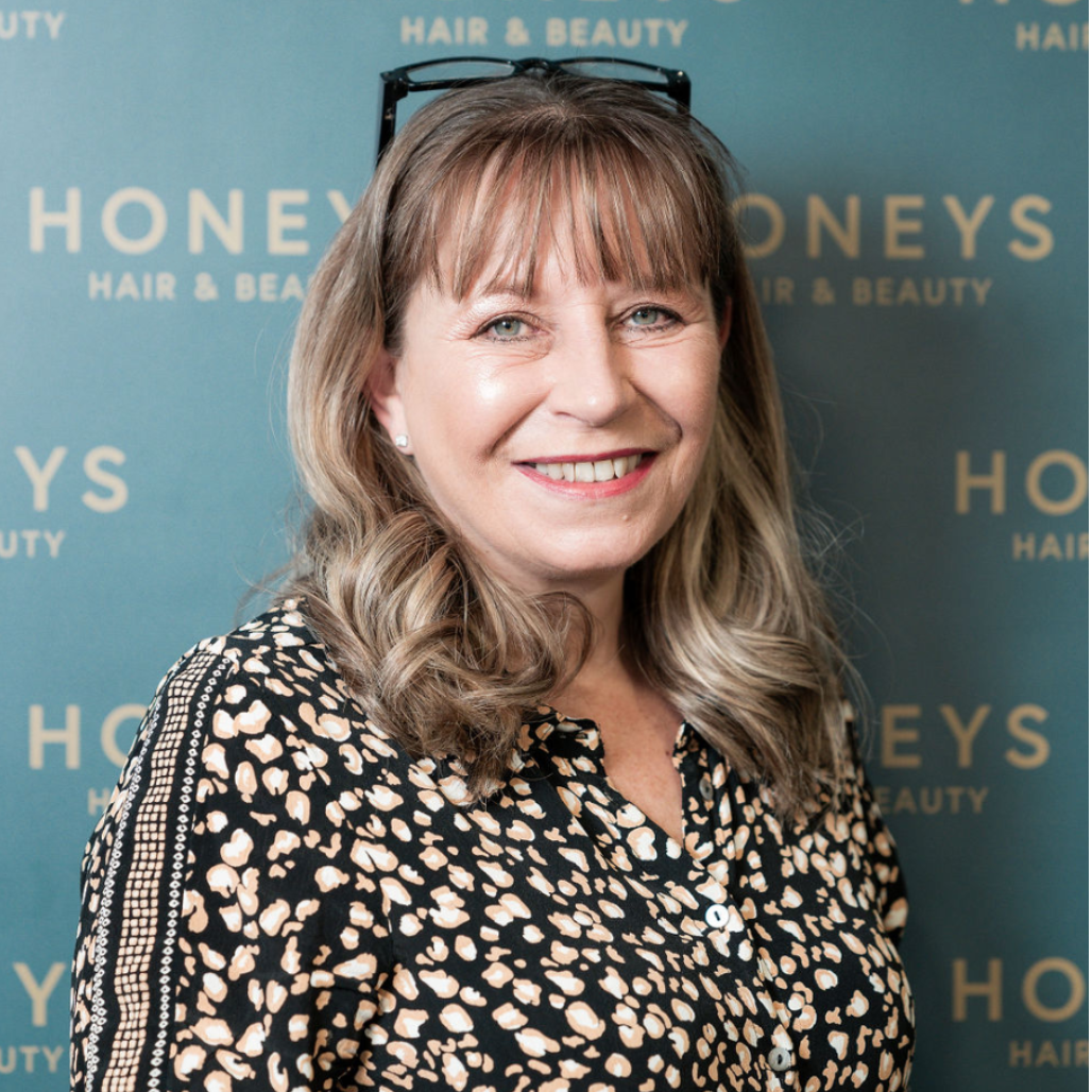 Sally Priscott, part of the Honeys Hair & Beauty team in Taunton, poses with a confident smile against the salon’s branded backdrop. With curled blonde hair and a chic printed blouse, she embodies professionalism and warmth.