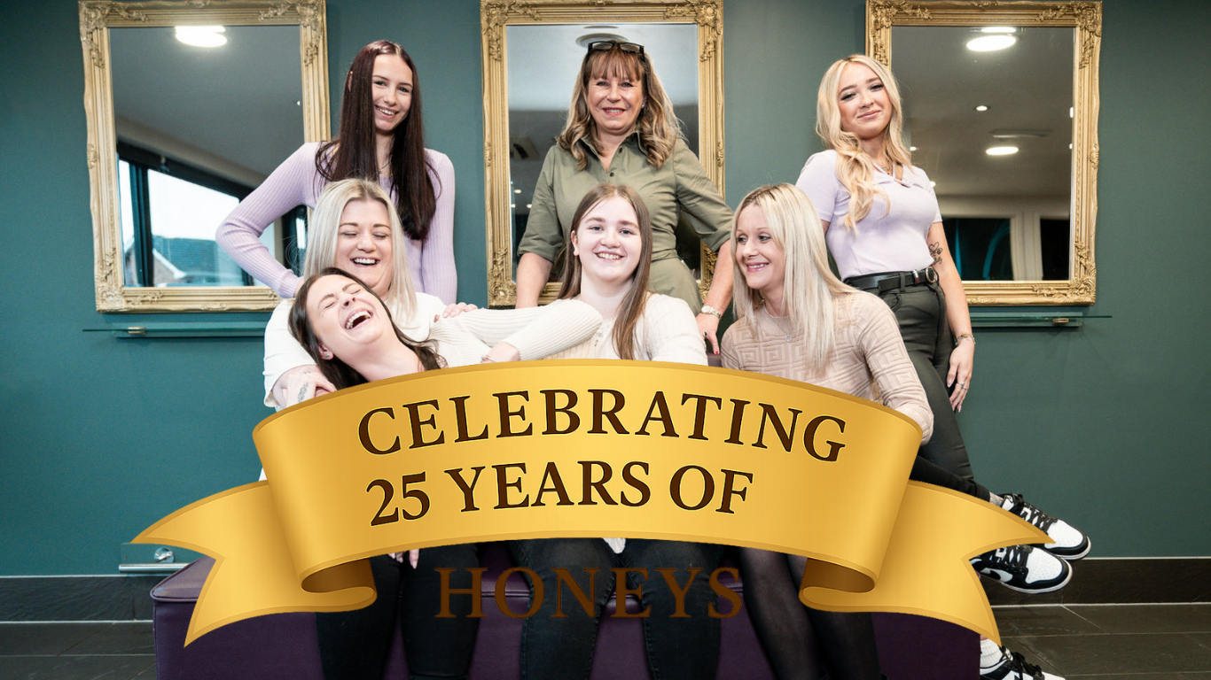 Seven smiling women pose in front of teal wall with gold-framed mirrors and a gold ribbon reading "CELEBRATING 25 YEARS OF HONEYS"