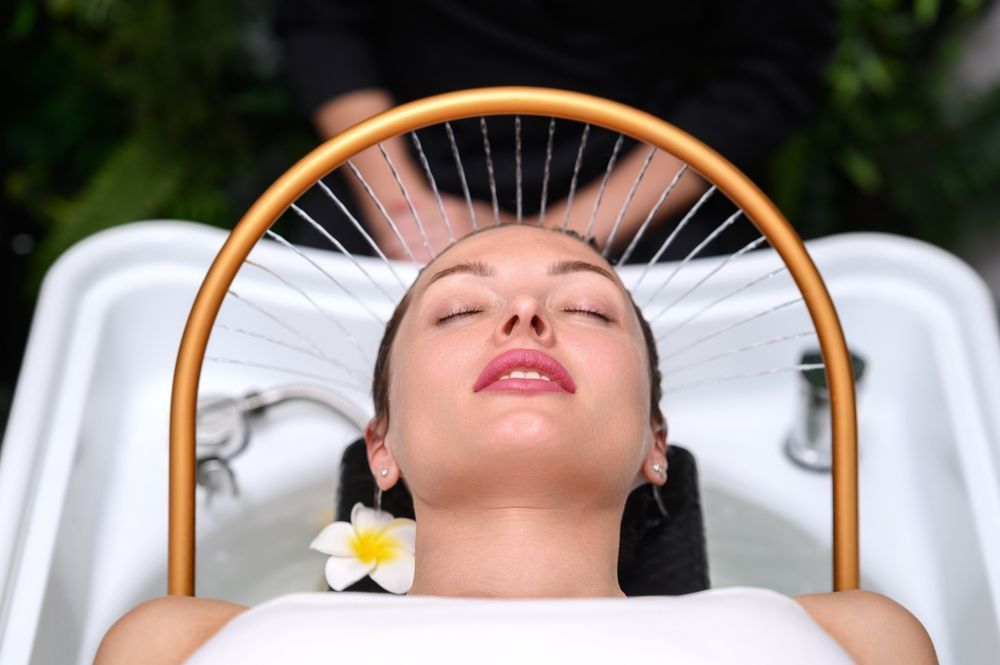 Spa client reclines at wash basin under halo-like head massage device; eyes closed, plumeria by ear