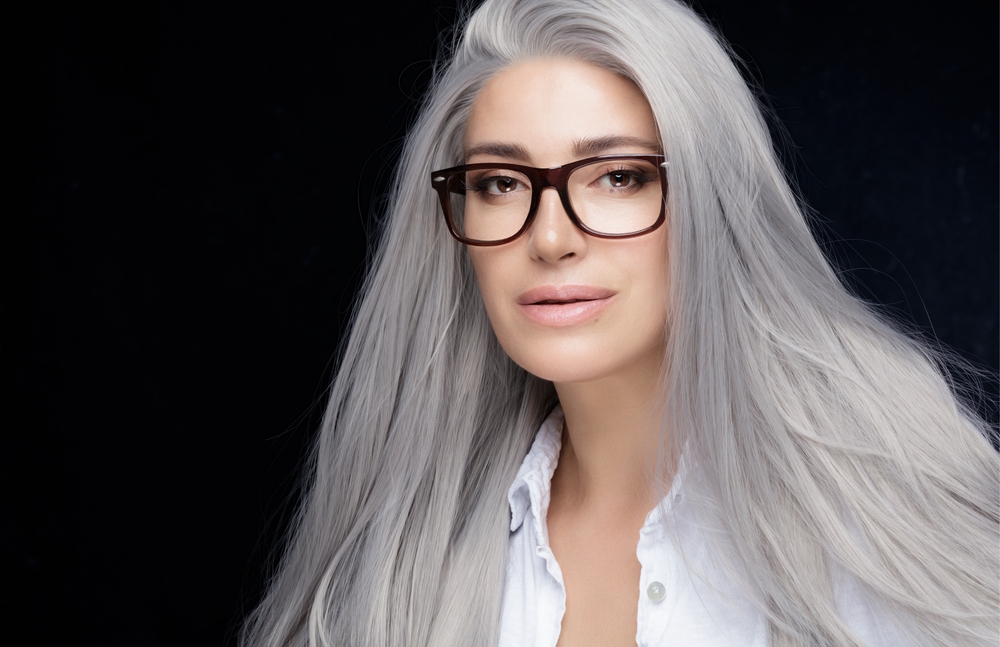 Woman with long silver-gray hair wearing dark-framed glasses and a white shirt against a dark background
